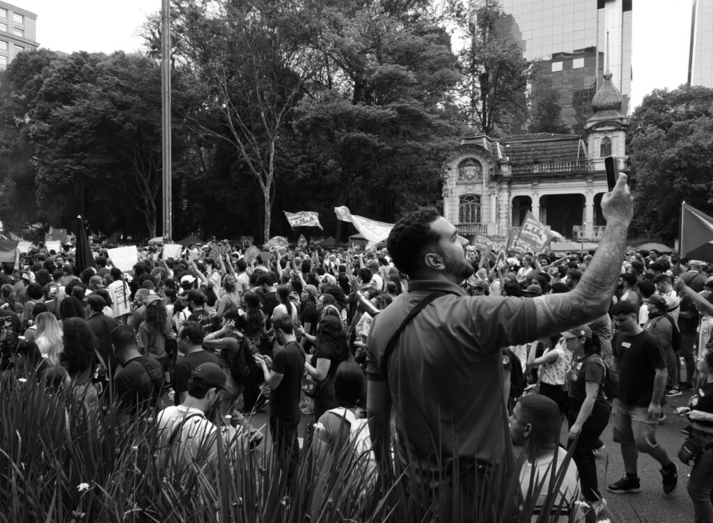 A man raises a mobile phone above a crowd during a public political demonstration on Avenida Paulista in São Paulo. The gesture records one observer making his own image within the larger gathering.