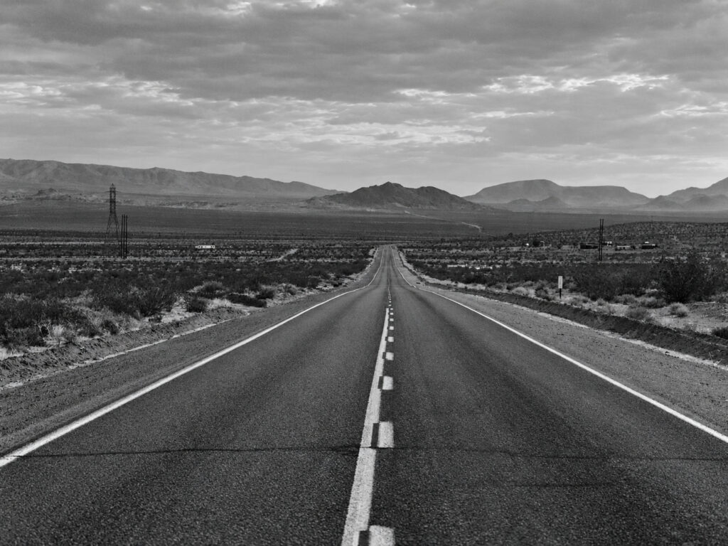 A straight highway leads through dry terrain towards distant mountains, offering a view of the open road shaped as much by engineering as by the idea of freedom.