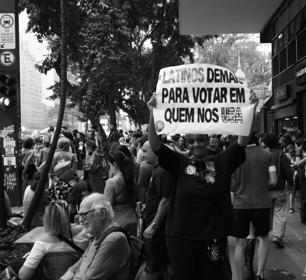 A demonstrator holds a handwritten sign above the surrounding crowd on Avenida Paulista. The photograph records the message as displayed, rather than making any claim about the opinions of others nearby.