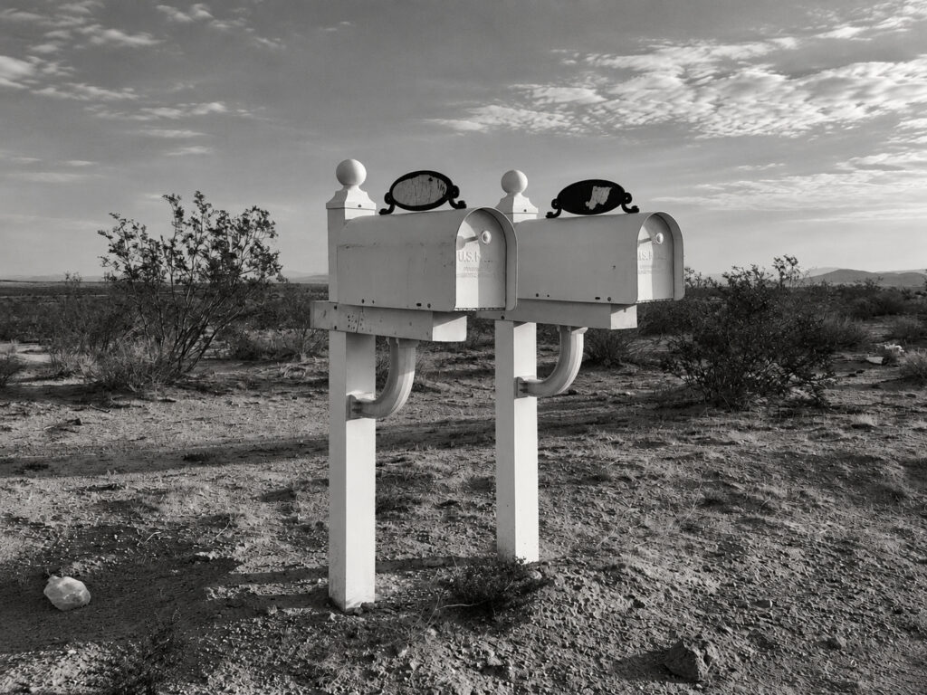 Two white roadside mailboxes stand beside one another, marking domestic addresses beyond the frame in a sparsely settled landscape.