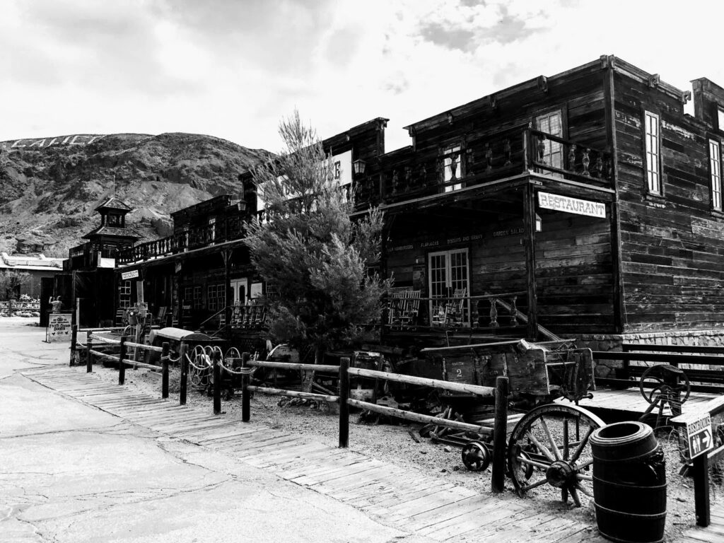 Wooden saloon and restaurant buildings line a street in Calico, with weathered boards, carts and wagon wheels reinforcing the town’s carefully maintained frontier appearance.