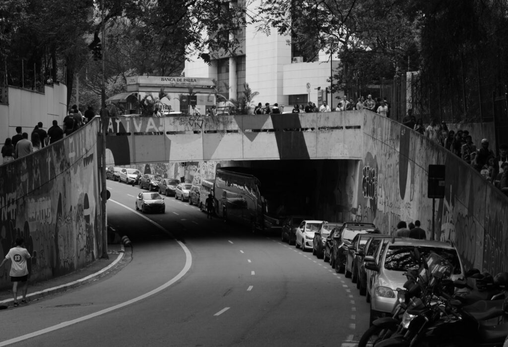 Black-and-white photograph: A quiet stretch of road curves beneath Avenida Paulista, framed by concrete walls, parked vehicles, and the city’s trees above