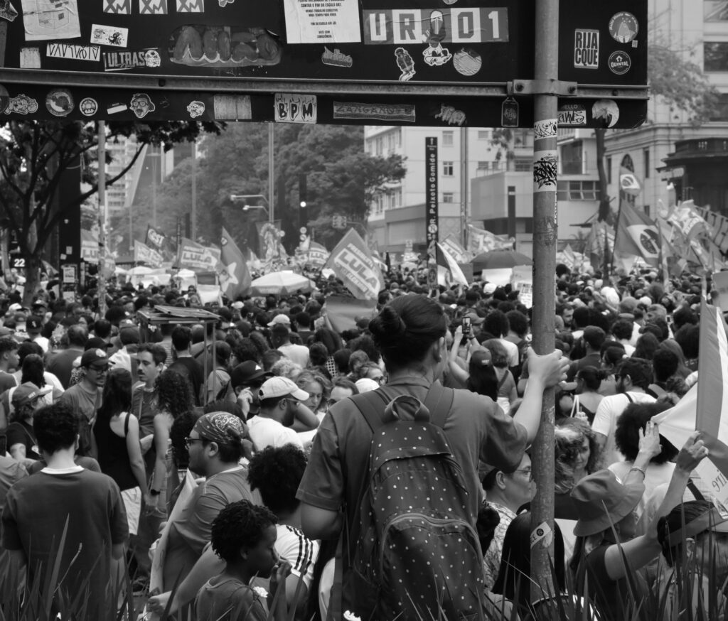 Black-and-white photograph: A dense crowd fills Avenida Paulista, with flags and banners rising above the gathering beneath a pedestrian overpass marked by the city