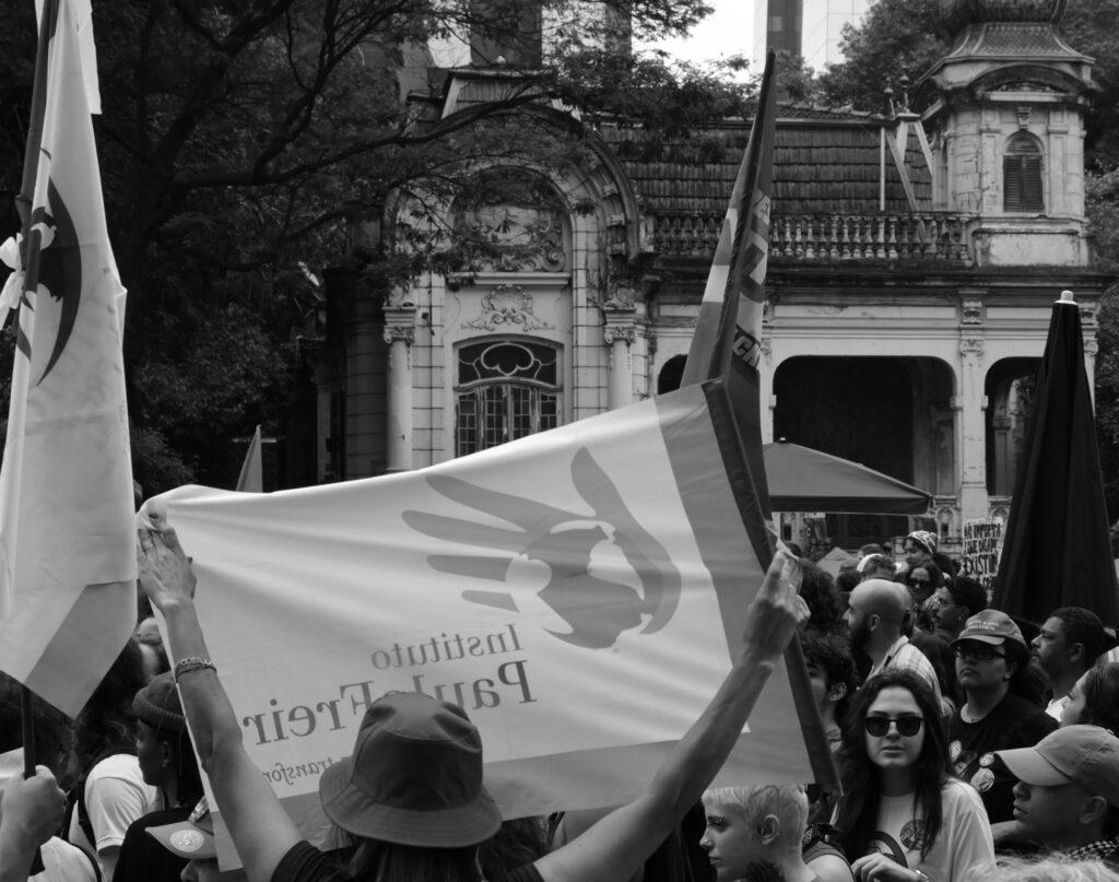 Black-and-white photograph: Political flags cross a crowd gathered in front of a historic building on Avenida Paulista