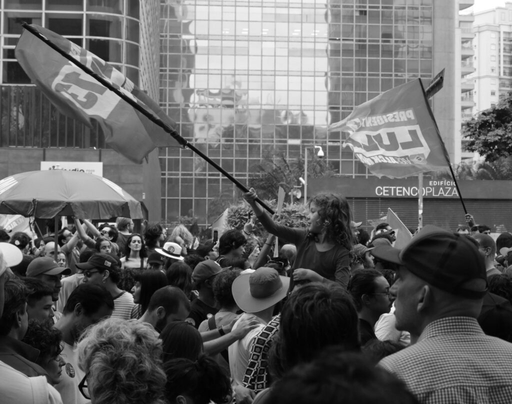 Black-and-white photograph: Flags cut diagonally across the frame as demonstrators gather shoulder to shoulder beneath the glass façades of São Paulo’s business district