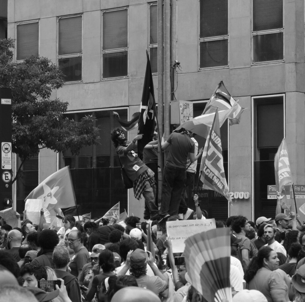 Black-and-white photograph: Above a tightly packed crowd, demonstrators raise flags against the geometric backdrop of an urban building