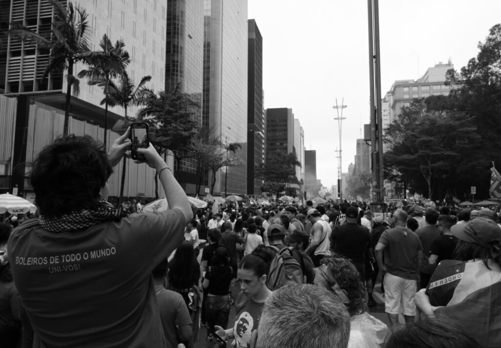 Black-and-white photograph: Seen from behind, a participant raises a phone to photograph the demonstration stretching along Avenida Paulista