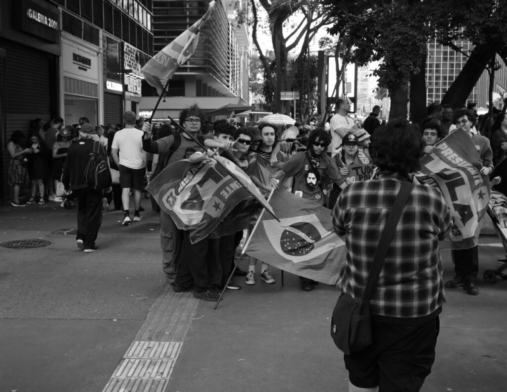 Black-and-white photograph: Political banners and flags converge on a tree-lined stretch of Avenida Paulista, carried by demonstrators moving along the busy sidewalk