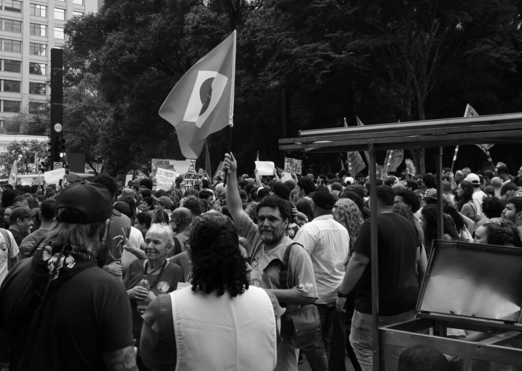 Black-and-white photograph: A crowd gathers beneath the trees of Avenida Paulista as a large flag rises above the people, its movement guiding the eye through the photograph