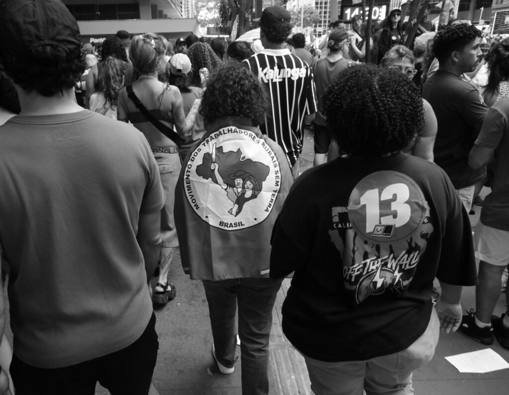 Black-and-white photograph: Photographed from behind, participants walk along a busy stretch of Avenida Paulista, wearing clothing printed with striking symbols and designs