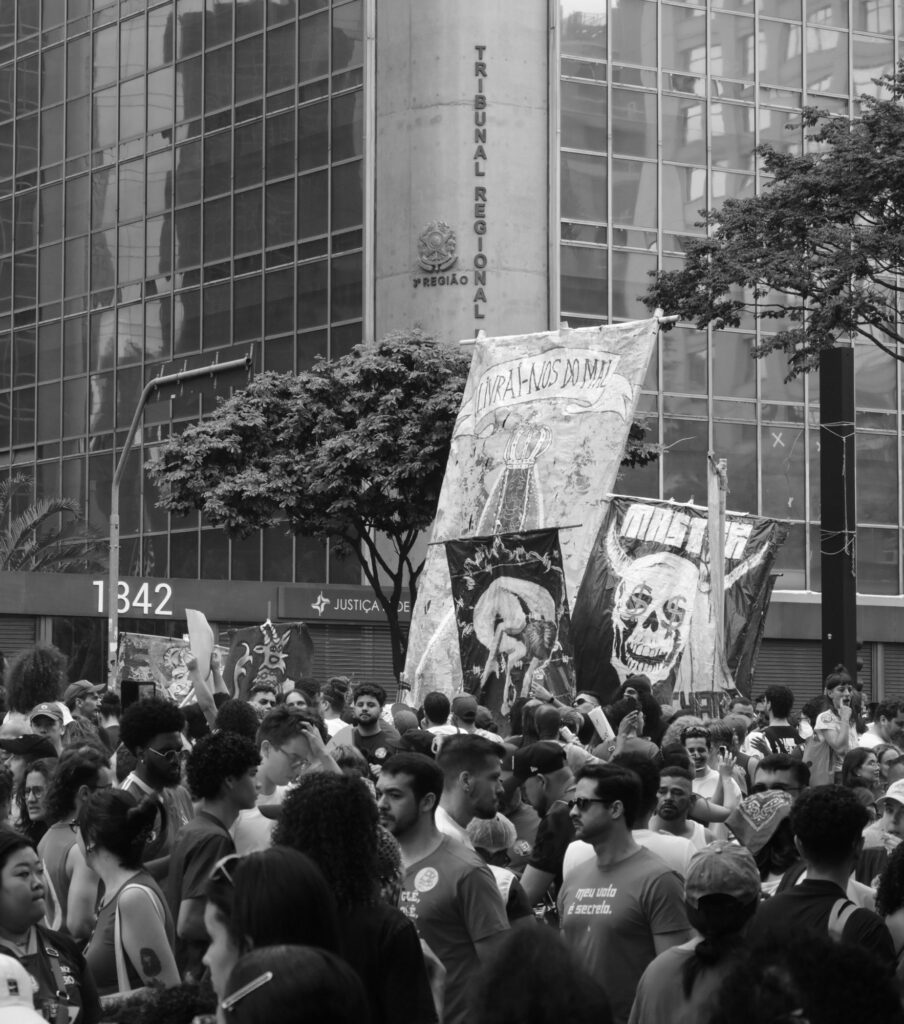Black-and-white photograph: Political banners rise above a dense crowd gathered between the glass towers and monumental façades of Avenida Paulista