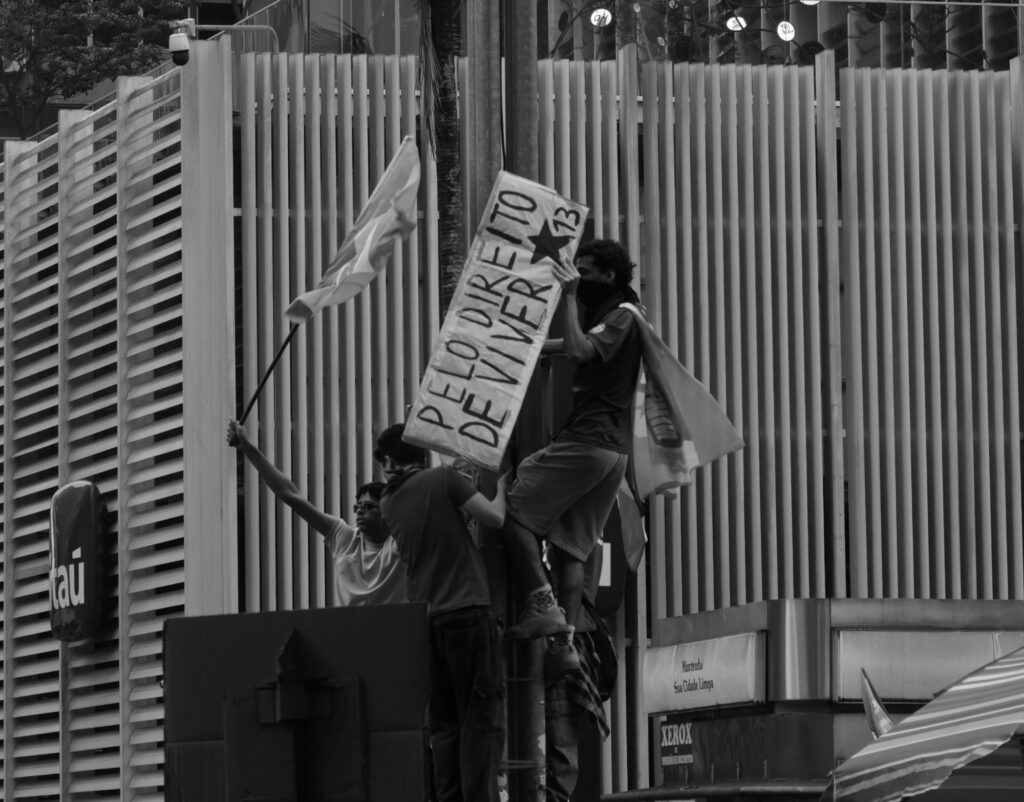 Black-and-white photograph: Two participants stand on an elevated structure in the street, holding flags and a political banner above the surrounding crowd