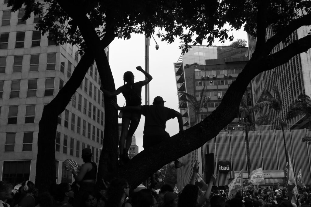 Black-and-white photograph: Silhouetted against the brightness of the city, several people occupy the branches of a large tree above the demonstration on Avenida Paulista