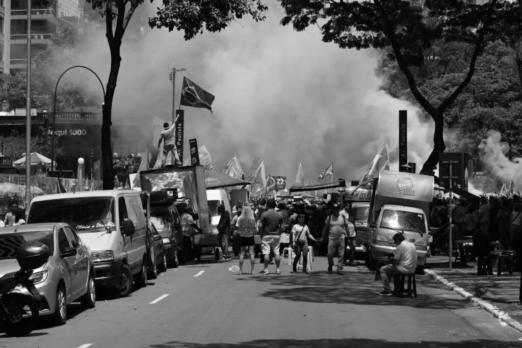 The gathering is seen from farther along the avenue, with smoke or haze visible in the distance. The viewpoint places the event within the broader streetscape rather than concentrating on the speakers or platform.
