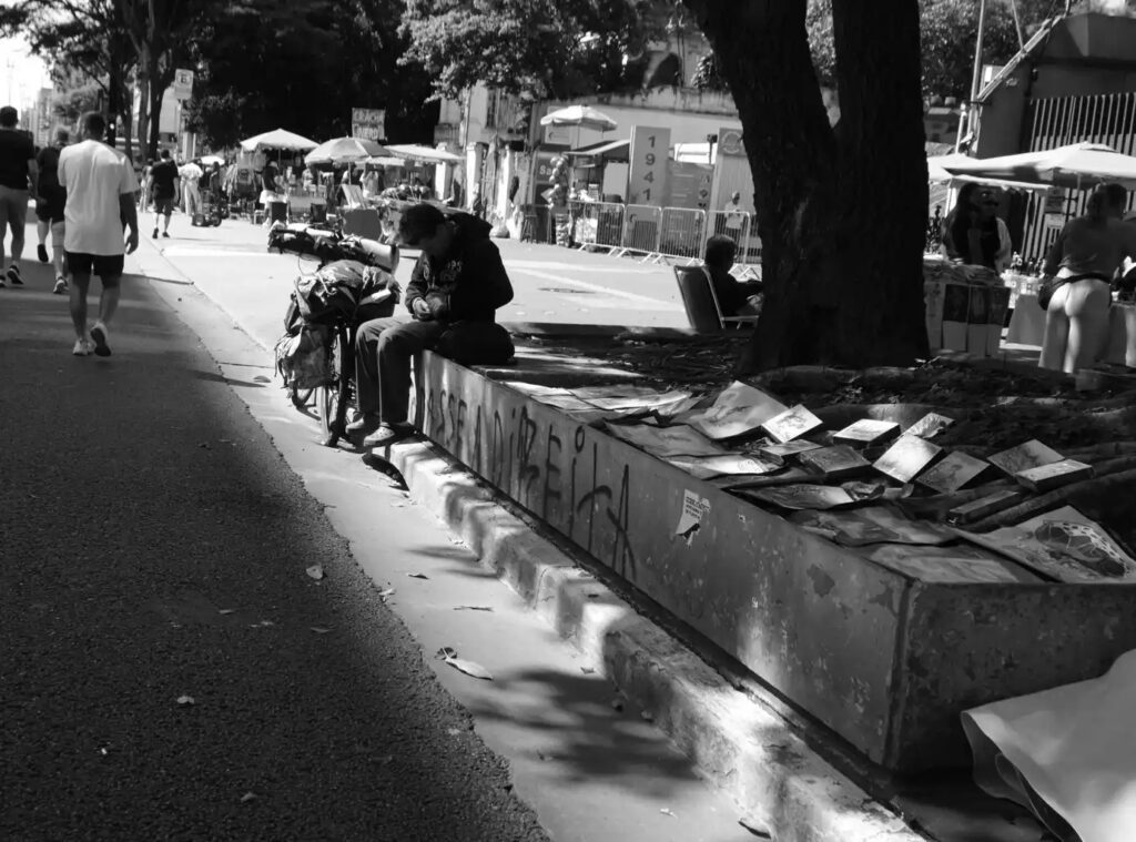 A shaded stretch of pavement contains people and everyday objects near the demonstration. The photograph redirects attention from the central gathering to the details of the surrounding street.