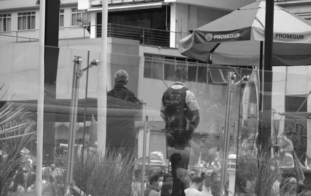 Figures and elements of the urban streetscape overlap within the frame. The photograph uses the surrounding structures and lines of sight to examine how people remain visible at the edge of the gathering.