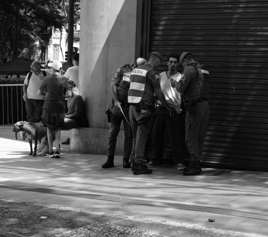 People occupy the pavement beside a closed shopfront. The shuttered frontage gives the photograph a quieter setting than the centre of the rally and draws attention to people whose activities are not explained by the public event.