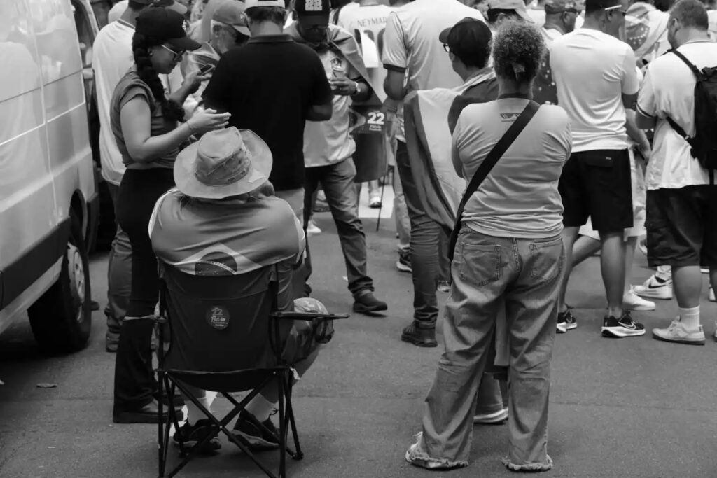 People stand and sit near the gathering, facing different directions and occupying the space in different ways. The photograph observes their presence without presuming what each person is waiting for or watching.