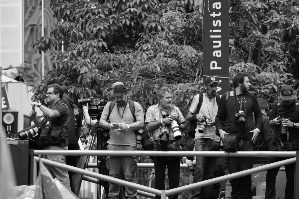 People watch activity on Avenida Paulista beneath or near a street sign. The photograph situates the onlookers within the recognisable urban setting of the demonstration.