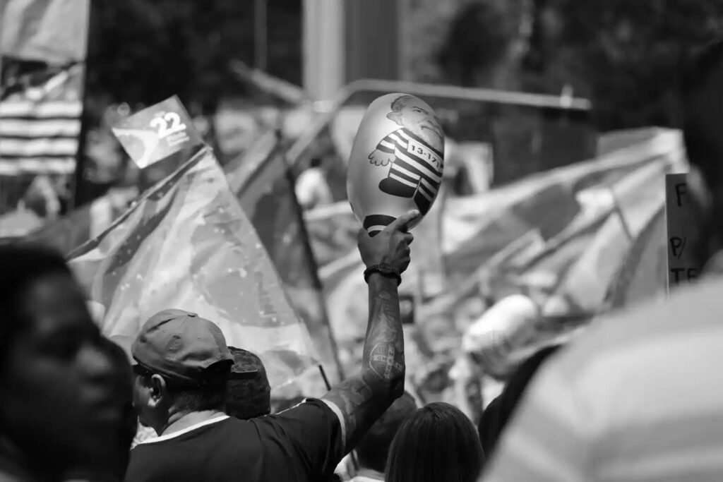 A political object is held above people gathered at the demonstration. The raised gesture draws attention within the crowd, while the rest of the frame places it among other participants and observers.