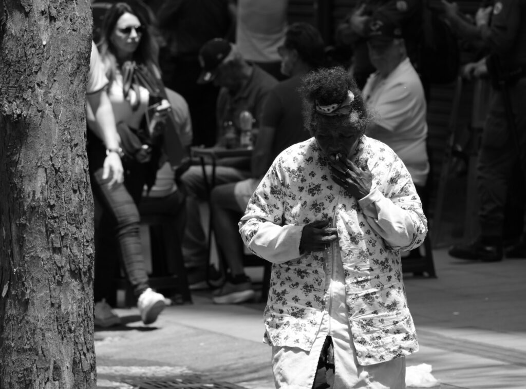 A person walks through a patch of sunlight near the gathering. The photograph closes the series with an ordinary movement through public space, away from the platform and the central political spectacle.