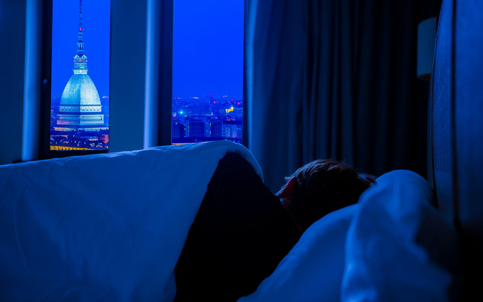A person asleep in a dark bedroom with a window looking out on a lit city skyline at night