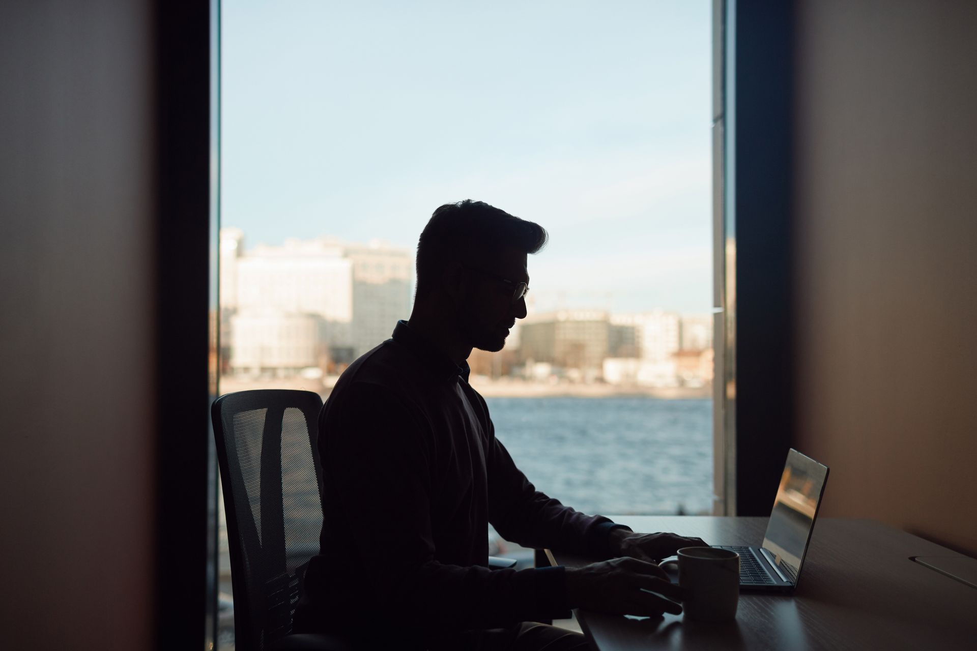 Silhouette of a person working from home on a laptop beside a large window